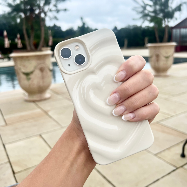 A hand with manicured nails holds the Melting Heart Phone Case - Coconut Cream by Coconut Lane, featuring a cream-colored, heart-patterned design. The background showcases an outdoor area with large potted plants, a swimming pool, and trees in the distance.