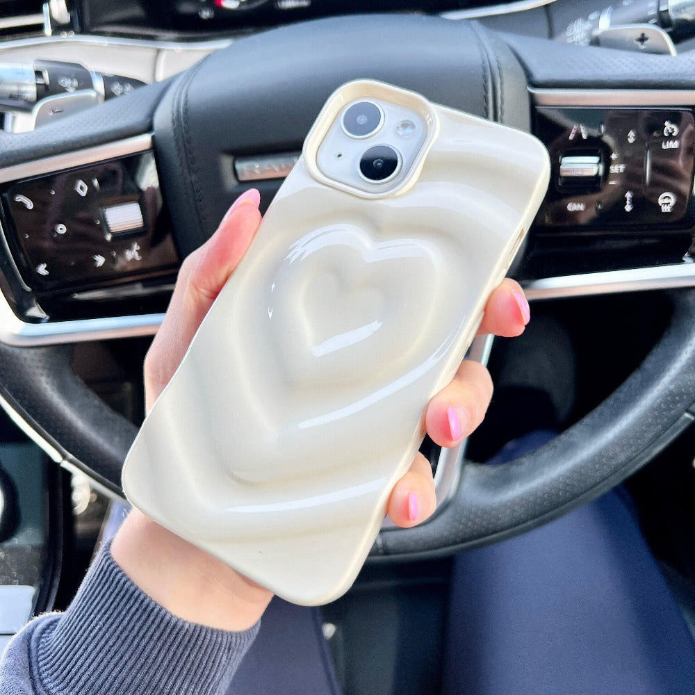 A hand with pink-painted nails holds a smartphone encased in the "Melting Heart Phone Case - Coconut Cream" by Coconut Lane. The light-colored, textured case features a raised heart design and is set against the backdrop of a car interior, with the steering wheel and dashboard controls clearly visible.