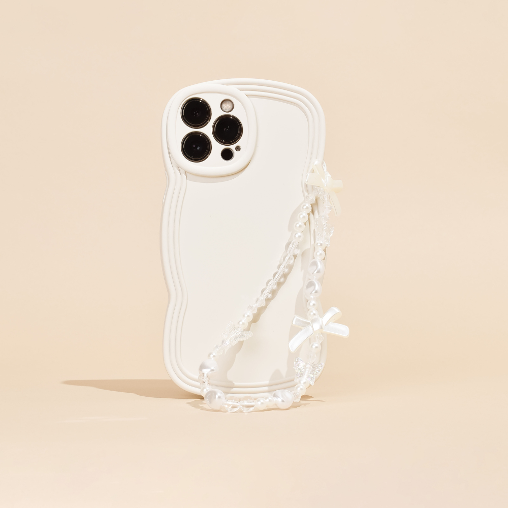 A white phone case with a wavy edge design is pictured against a beige background. This chic phone accessory from Coconut Lane features three camera cutouts and the Beaded Phone Strap - Bows & Butterflies, a decorative pearl chain with small white bows and butterflies attached, hanging from one side. The beaded phone strap adds an extra touch of elegance.