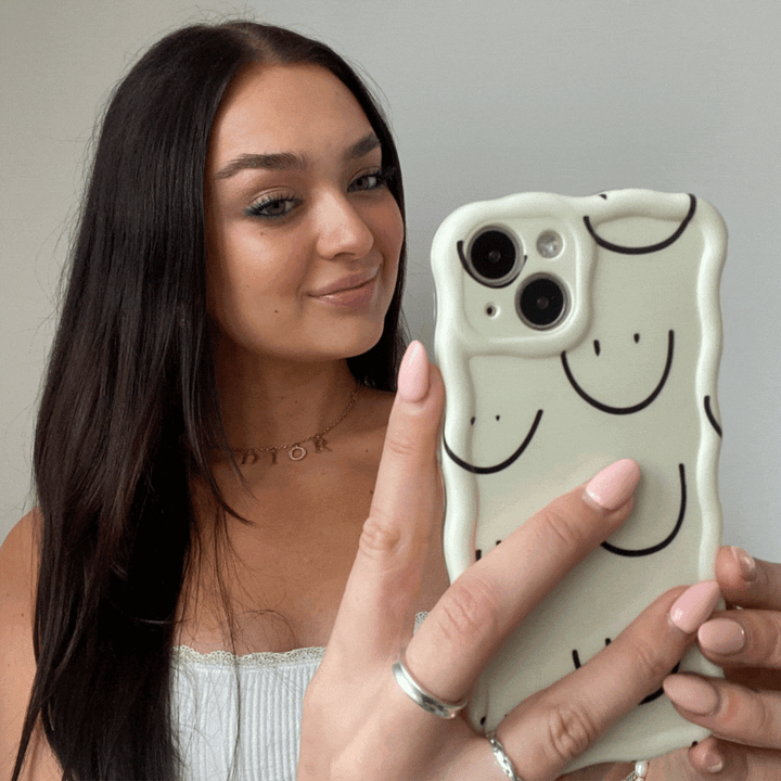 A person with long dark hair, wearing a light-colored top, is taking a selfie with a smartphone. The extra protective phone case is the Wavy Phone Case - Smiley Doodle by Coconut Lane, featuring black smiley doodle designs on a white background. The person is holding the phone in their left hand and has multiple rings on their fingers.