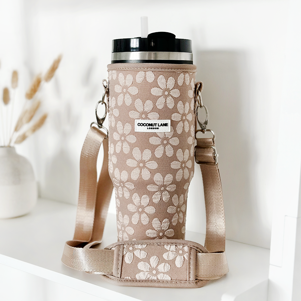 A beige insulated Tumbler Carry Case, named "Mocha Flower" by Coconut Lane, adorned with a floral pattern, features a handle and strap for portability, and comes with a black lid accompanied by a white straw. It is labeled "Coconut Lane" on the front and is placed on a white surface with dried flowers in the background.
