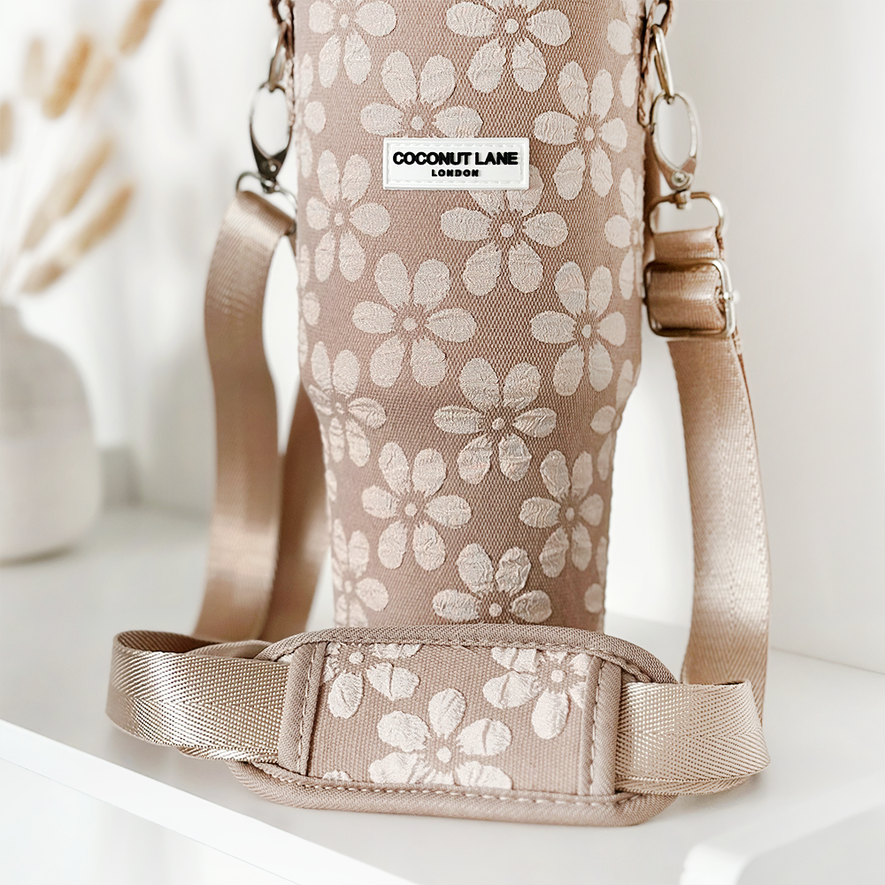 A bag named "Tumbler Carry Case - Mocha Flower" with a beige base and floral pattern, featuring adjustable straps, is displayed as the perfect companion for your Stanley Quencher. The brand label reads "Coconut Lane." Positioned against a white background, blurred decor items like dried flowers in a vase add charm in the distance.

