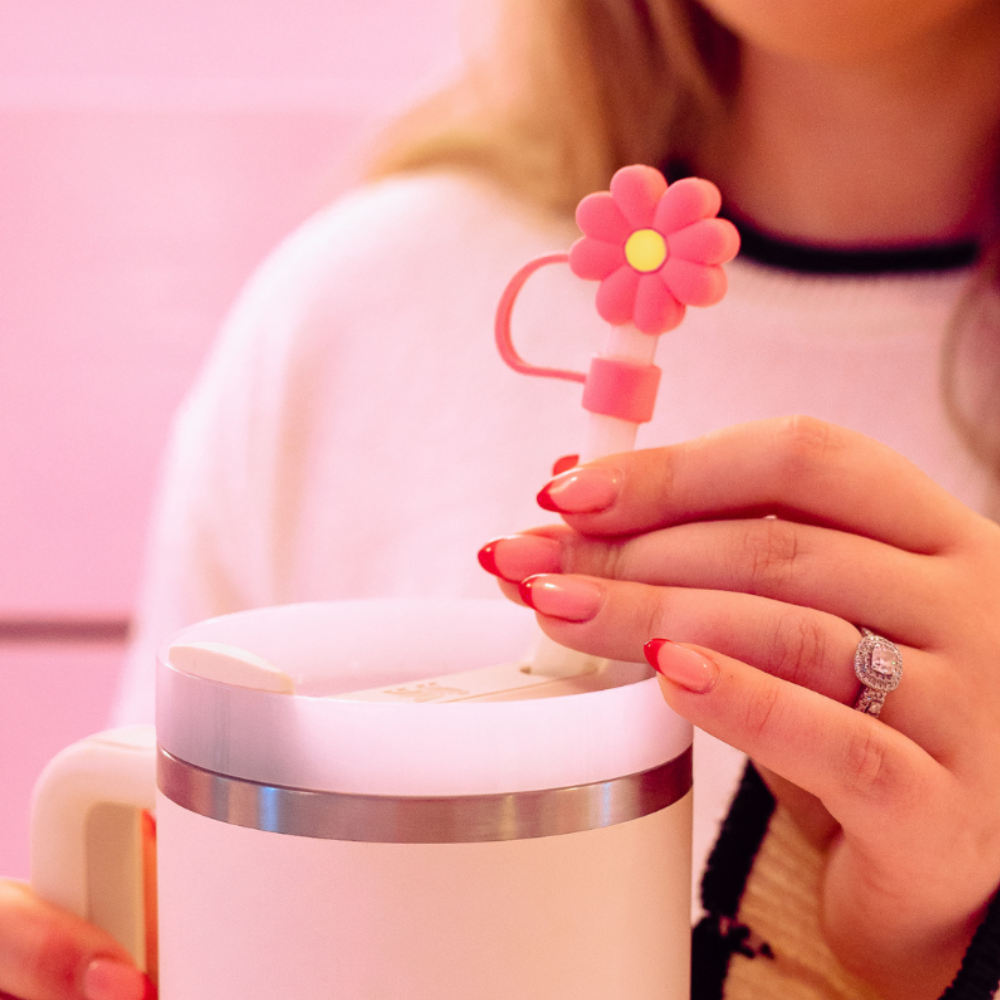 A person with long, manicured nails and a diamond ring holds a small Pink & Yellow Daisy Straw Cover by Coconut Lane above a white insulated mug. The background is pink, and the person is dressed in a light-colored sweater.