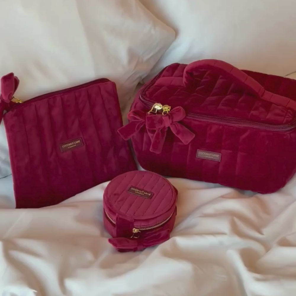 Burgundy velvet cosmetic bag, jewellery box and pouch with a quilted design, top handle, bow detail, and gold-tone hardware. Sit on a white bed