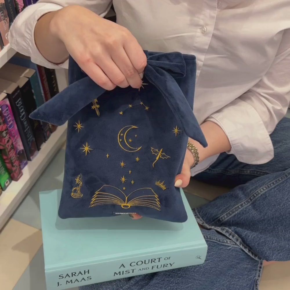 Book Sleeve made from soft navy blue velvet with gold embroidered crescent moon, stars, crown and open book illustrations, finished with a large velvet bow and Coconut Lane logo, photographed on a white background.