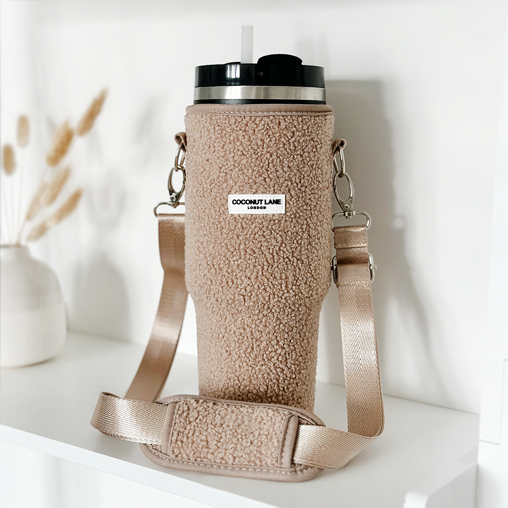 A beige tumbler with a fuzzy, textured cover called the "Tumbler Carry Case - Teddy Rupert" from Coconut Lane, featuring a convenient strap for carrying. The label on the cover reads "Coconut Lane." It sits on a white shelf near a white vase with dried grasses, blending a cozy and minimalist aesthetic. This travel mug ensures stylish tumbler protection wherever you go.
