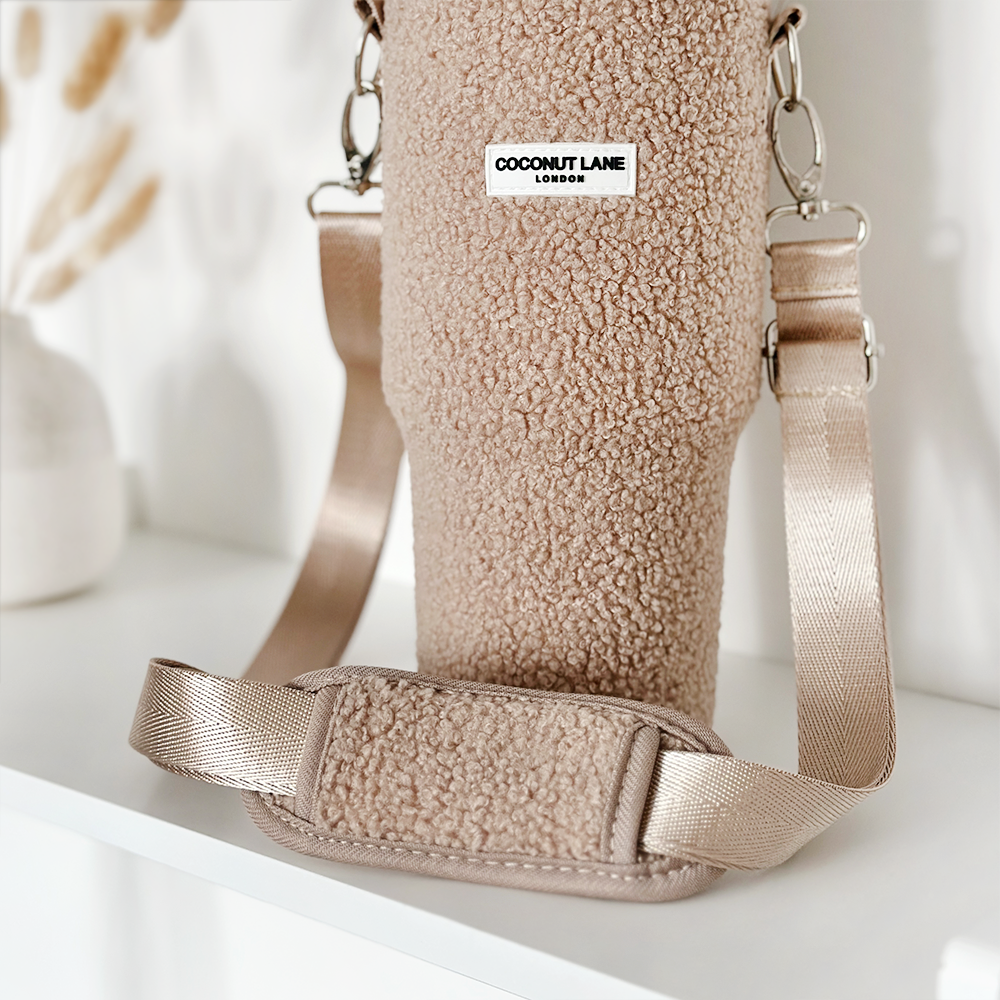 A Teddy Rupert Tumbler Carry Case from Coconut Lane, featuring a metallic beige strap, sits on a white shelf. This fuzzy beige holder is ideal for travel mug protection and has a label reading "Coconut Lane London." The background showcases a blurred white vase with dried beige plants.