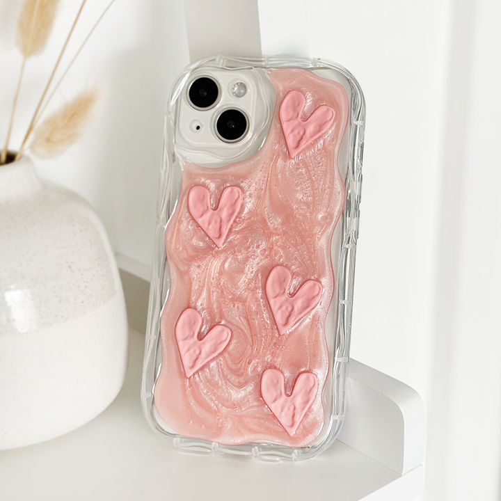 A Coconut Lane Shimmer Pink Hearts Phone Case featuring an extra protective clear design with pink swirls and five raised shimmer pink hearts is propped up against a white vase with dried wheat stalks. The background is a clean, white surface.