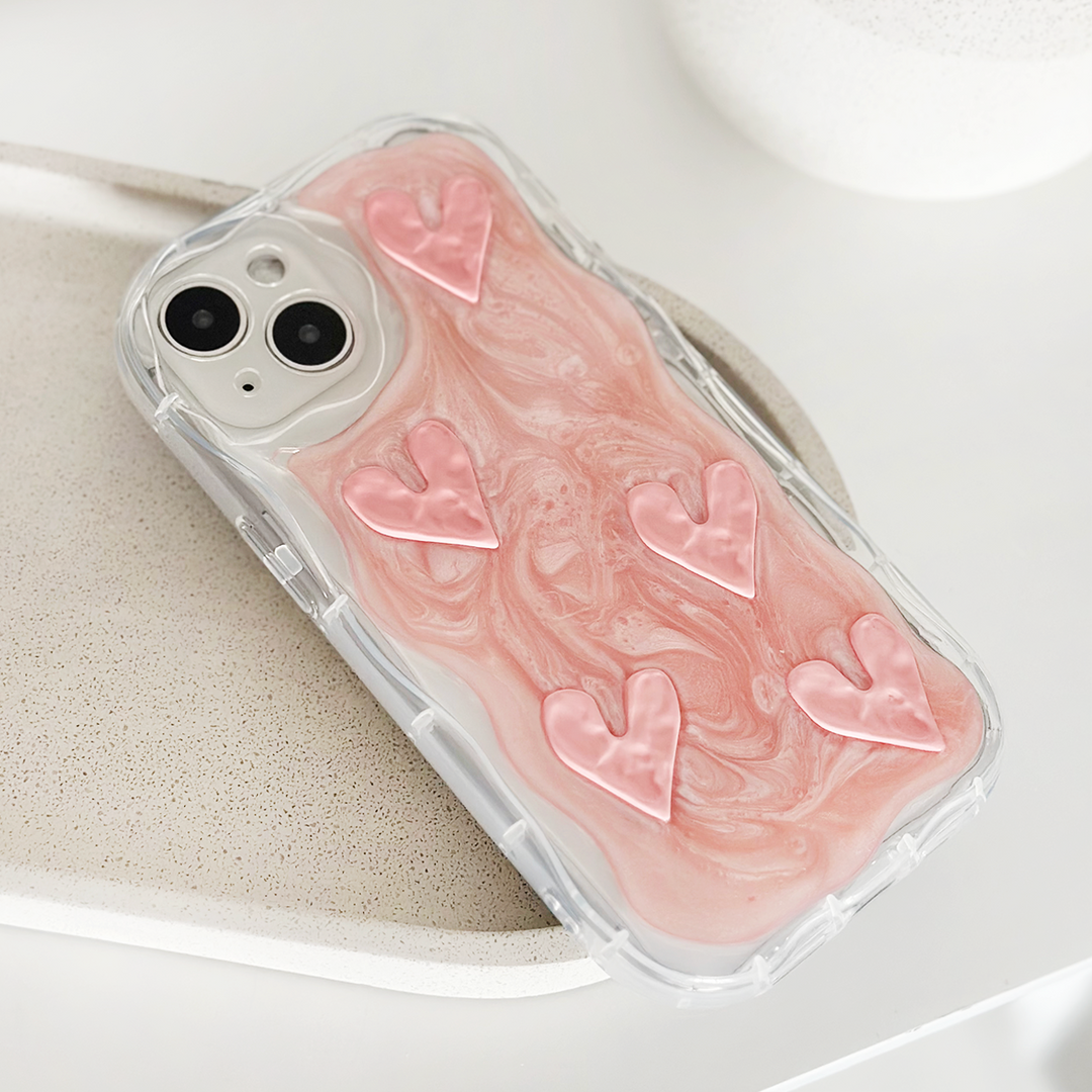 A smartphone encased in Coconut Lane's Shimmer Pink Hearts Phone Case, an extra protective transparent cover showcasing a pink and white marbled design adorned with six raised shimmer pink hearts. The phone is set on a light-colored speckled surface, creating a clean and minimalist aesthetic.