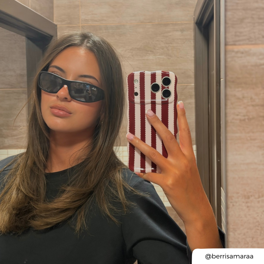 Woman with brunette hair holds up iphone taking a mirror selfie. The phone is in a burgundy and light pink striped wavy phone case.