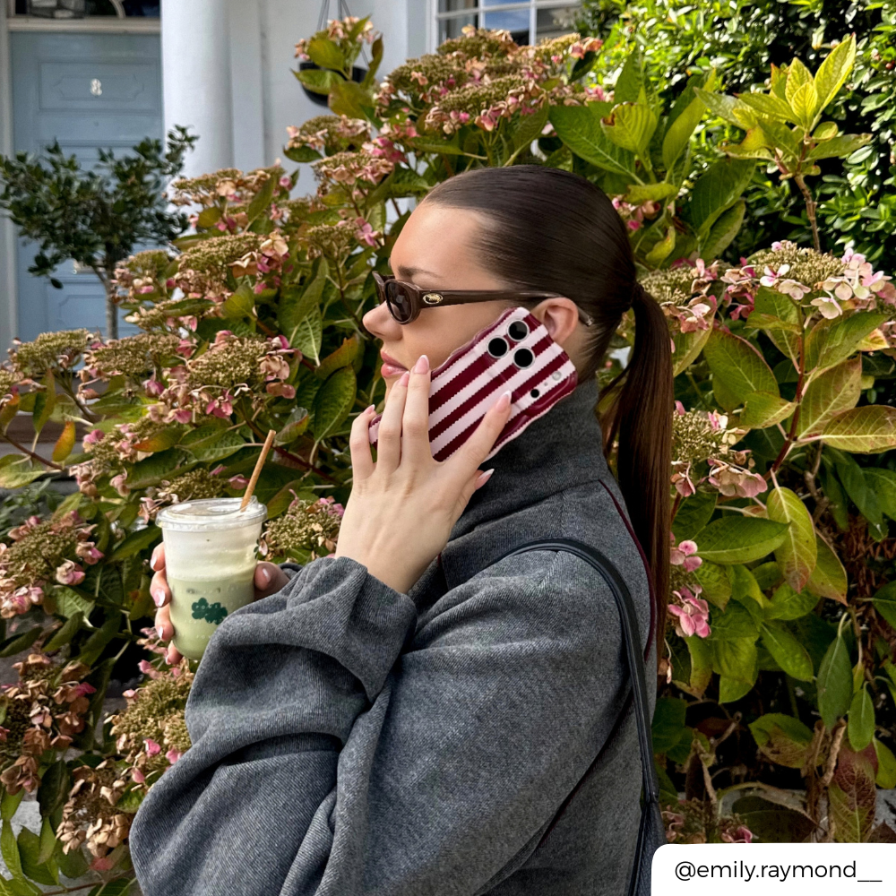 Woman with brown hair holds up iphone to her ear. The phone is in a burgundy and light pink striped wavy phone case.