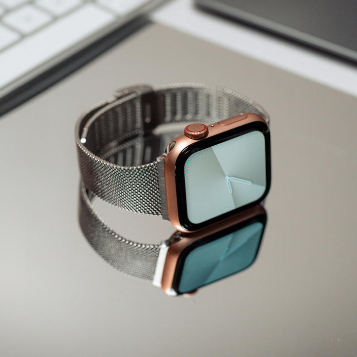 A smartwatch featuring the Coconut Lane Milanese Apple Watch Strap - Silver lies on a reflective surface. The watch face displays an analog clock with simple white hands. Positioned near a keyboard and a laptop, with their edges partially visible in the background, this strap is compatible with Apple Watch models.