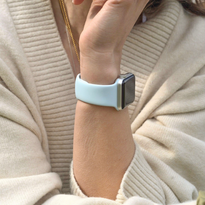 A person wearing a cream-colored sweater rests their chin on their hand, showcasing a Coconut Lane NAKD Apple Watch Strap with a sky blue silicone band. Complementing the look is a thin gold necklace, bringing an elegant touch to the spring-inspired pastels ensemble.