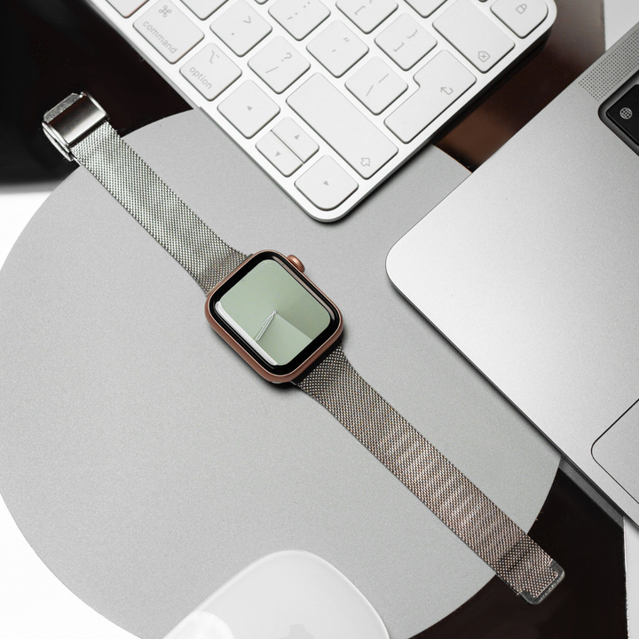 A modern and minimalistic scene features a smartwatch with a Milanese Apple Watch Strap - Silver by Coconut Lane placed on a surface next to a keyboard, part of a laptop, and other electronic devices. The smartwatch screen displays an analog clock face.