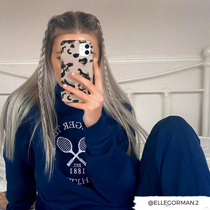 A person with long, light-colored hair styled in braids is sitting in bed while taking a mirror selfie with a phone encased in Coconut Lane's Ivory Tort Phone Case. The person is wearing a dark-colored sweatshirt featuring a tennis-themed design. The Instagram handle "@ELLEGORMAN.2" is visible on the image.