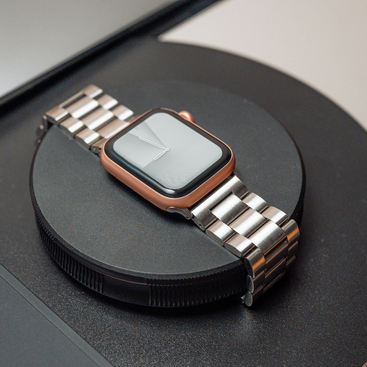 A smartwatch with a rectangular screen and metal band rests on a round black wireless charging pad. The watch, featuring a rose gold bezel, is turned off with its screen blank. An adjustable strap made of Stainless Steel Apple Watch Strap - Silver from Coconut Lane complements the sleek design. The background shows a blurred surface and part of another device.