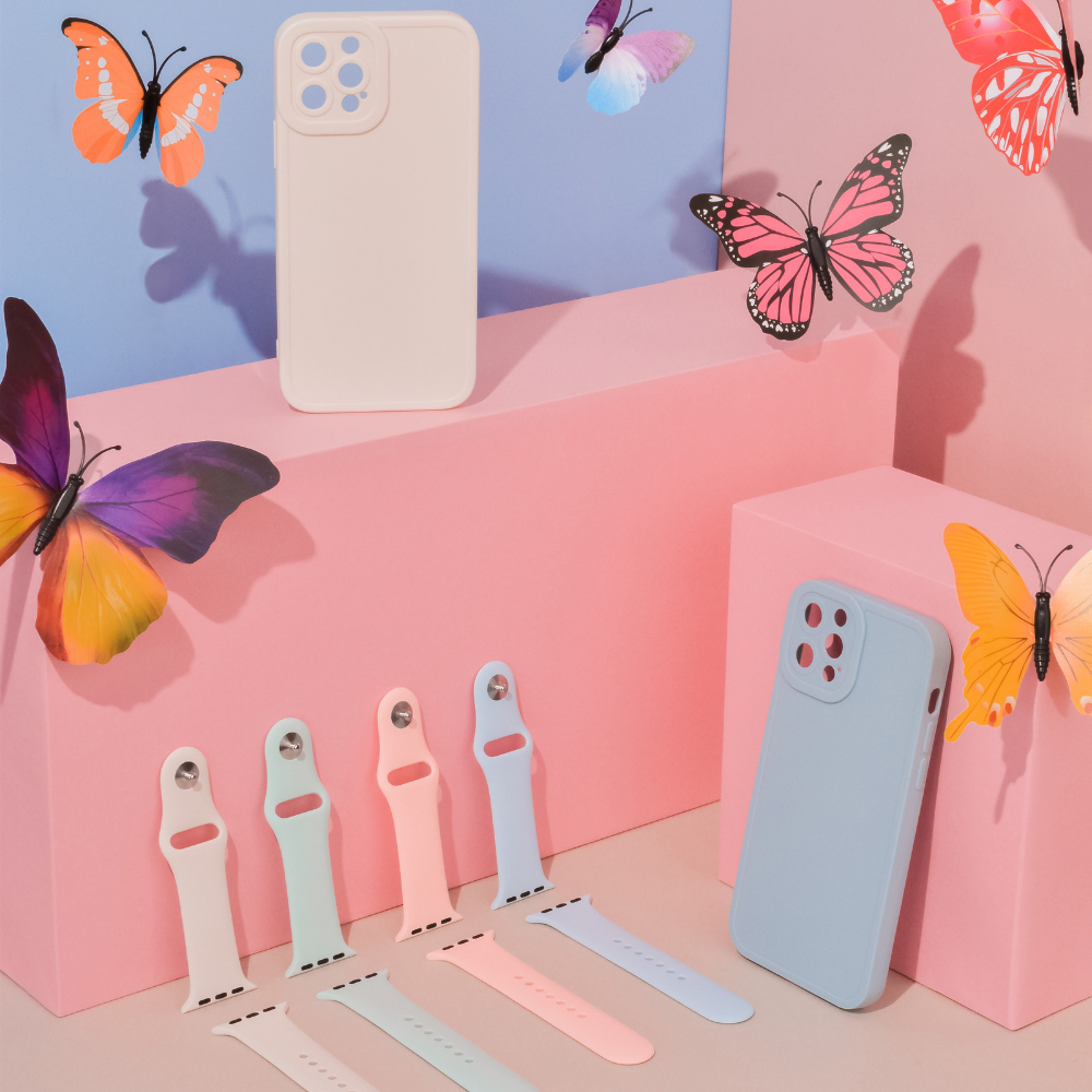 A display of colorful phone accessories against a pastel background showcases several phone cases and watch bands in pink, blue, green, and white shades. Among them, the NAKD Apple Watch Strap in Sky Blue by Coconut Lane blends seamlessly into the scene with spring-inspired pastels, enhanced by vibrant butterflies.