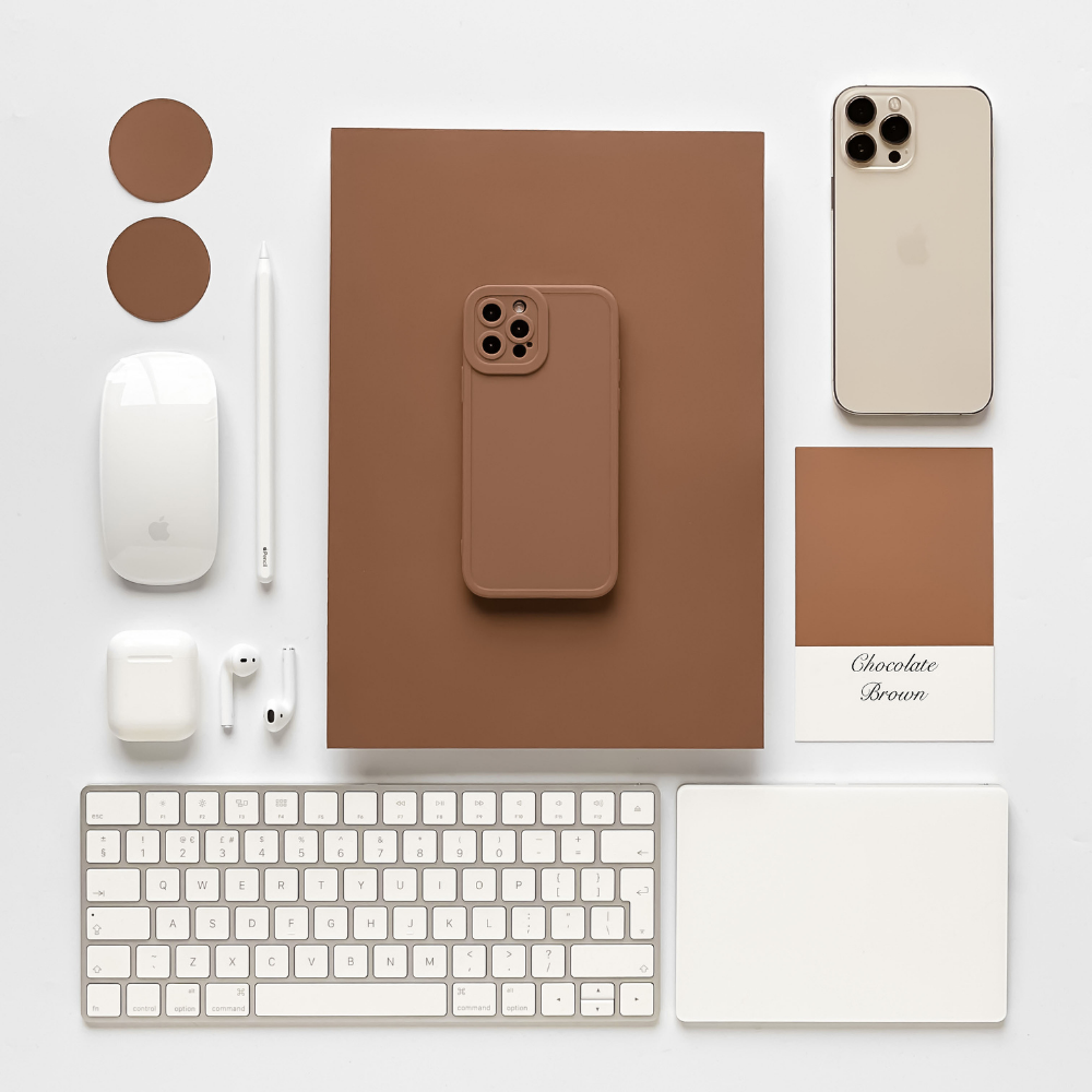 An overhead view of a neatly arranged workspace featuring a brown notebook, a NAKD Phone Case - Chocolate Brown by Coconut Lane, a silver iPhone, an Apple Magic Mouse, an Apple Magic Keyboard, AirPods, a white stylus, and two brown coasters. A color swatch labeled "Chocolate Brown" complements the NAKD Phone Case - Chocolate Brown on display.