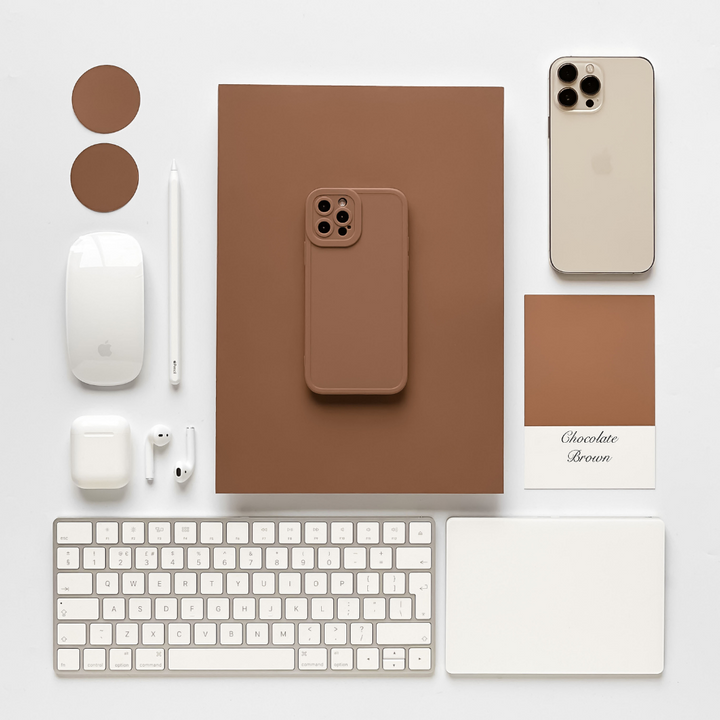 An overhead view of a neatly arranged workspace featuring a brown notebook, a NAKD Phone Case - Chocolate Brown by Coconut Lane, a silver iPhone, an Apple Magic Mouse, an Apple Magic Keyboard, AirPods, a white stylus, and two brown coasters. A color swatch labeled "Chocolate Brown" complements the NAKD Phone Case - Chocolate Brown on display.
