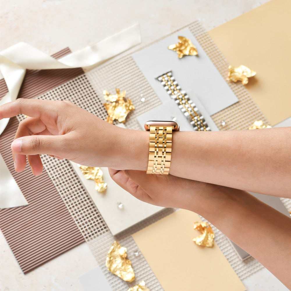 A person wearing the Stainless Steel Apple Watch Strap - Gold by Coconut Lane is adjusting the strap on their wrist. The background features decorative elements like golden foil accents, ribbons, and textured papers in neutral tones.