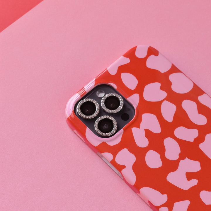 A smartphone encased in a protective cover featuring a red and pink cow print pattern, with Coconut Lane's Diamante Protective Camera Lens Covers enhancing the iPhone’s lenses. These covers include a decorative ring of small, sparkling stones and scratch-resistant tempered glass. The background is made up of various shades of pink paper.