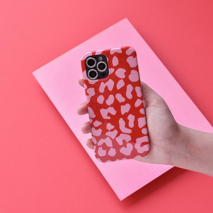 A hand holding an iPhone with a red and pink patterned case against a pink and red background. The phone's camera lenses are visible, protected by Coconut Lane's tempered glass "Protective Camera Lens Covers - Diamante," while the scratch-resistant case features abstract heart-like shapes.