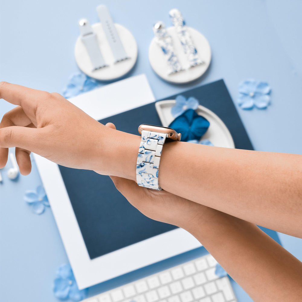A person wearing a Coconut Lane Luxe Blue & White Apple Watch Strap is wrist-deep over a light blue table. In the background, various blue and white objects, including an adjustable Apple Watch strap, watch bands, a keyboard, and flower-shaped decorations, are neatly arranged on the table.