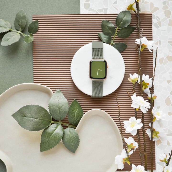 The Coconut Lane Magnetic Chain Apple Watch Strap in Khaki is showcased elegantly on a white round stand, encircled by various plants and flowers. This stylish wristwatch strap, complete with a gold-colored case and green strap, is set against a backdrop featuring green, brown, and white mosaic patterns that subtly enhance its design. It fits all Apple Watch models.