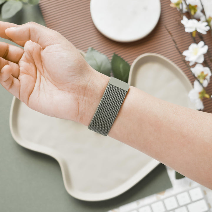 A close-up of a wrist wearing the Coconut Lane Magnetic Chain Apple Watch Strap - Khaki, featuring a sleek, minimalist design with a Stainless Steel Magnetic Clasp. The background displays a decorative arrangement including a round white dish, white flowers, green leaves, and part of a keyboard, all set on a green surface.