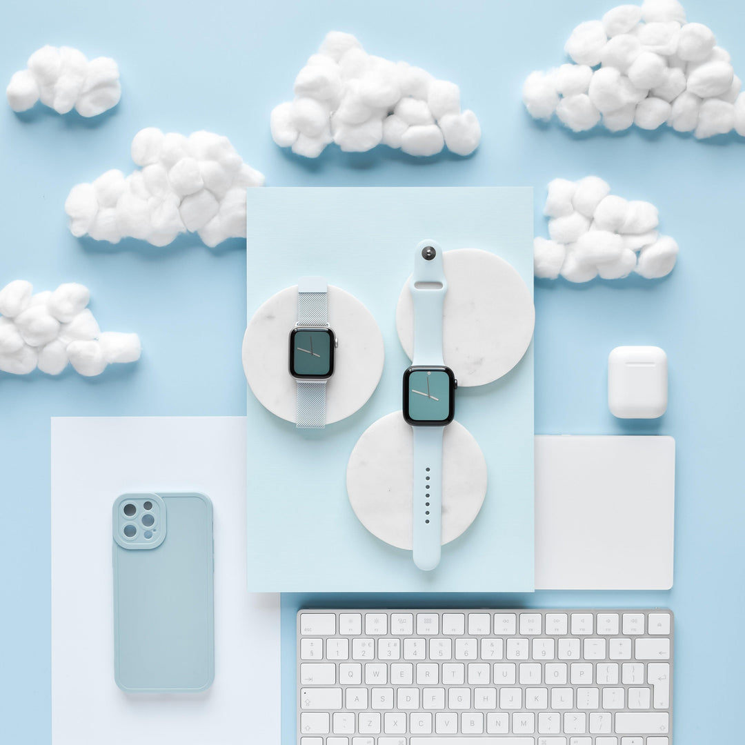 A flat lay of smartwatch and tech accessories includes two smartwatches with pastel bands on marble coasters, a Coconut Lane NAKD Phone Case in baby blue featuring soft flexible TPU for full camera protection, a white keyboard, a computer mouse, and an AirPods case. The background is light blue with fluffy white cotton clouds.