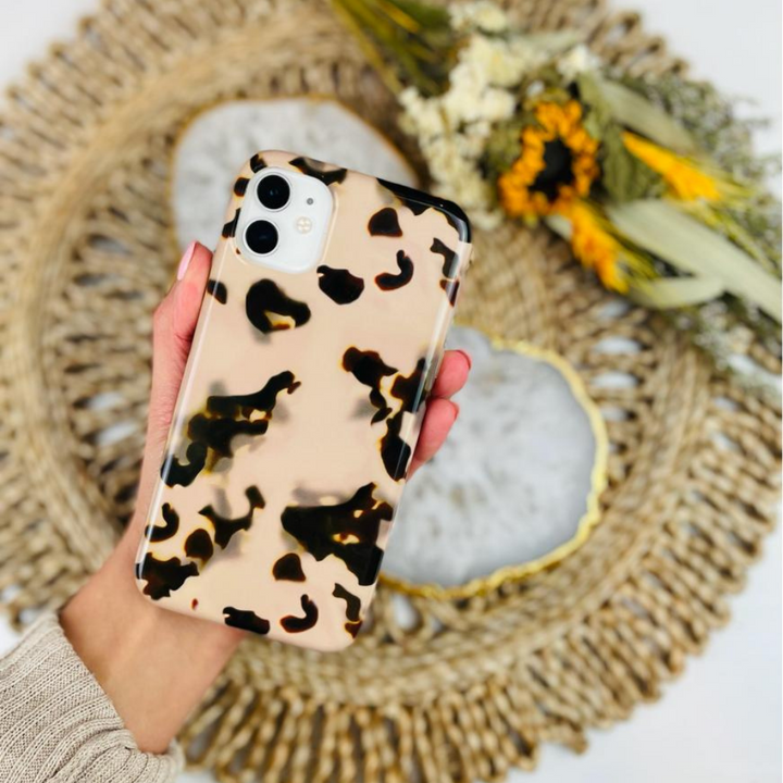 A hand holds an iPhone adorned with the Ivory Tort Phone Case by Coconut Lane, featuring a stylish beige and black tortoiseshell pattern. In the background, a rustic woven surface enhances a decorative centerpiece that includes a white stone slab with gold edging and dried sunflowers. This stunning Ivory Tort Phone Case from Coconut Lane adds a touch of elegance to your style.