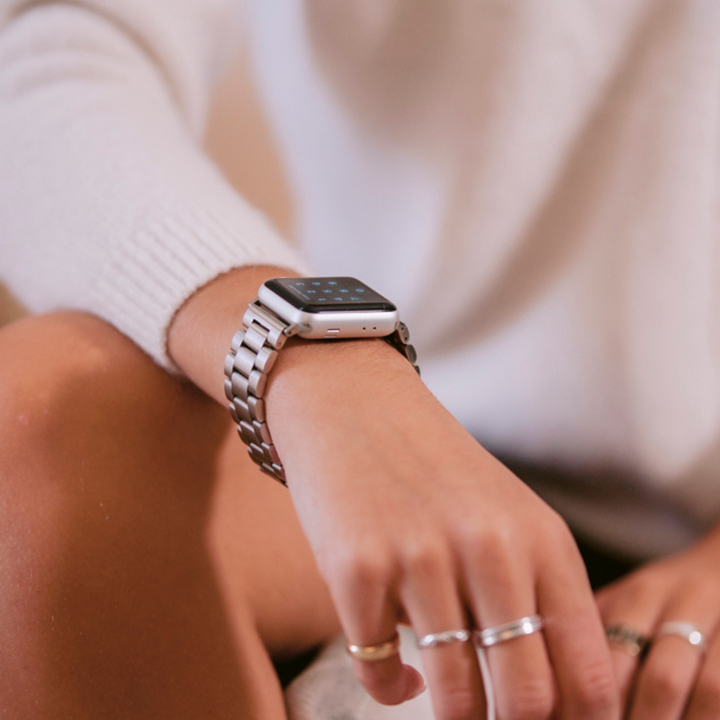 Close-up of a person wearing a smartwatch with the Stainless Steel Apple Watch Strap - Silver by Coconut Lane on their left wrist. They are also wearing multiple rings on their fingers. The person is dressed in a white long-sleeve shirt, and their arm, adorned with the sleek Coconut Lane strap, is resting on their knee.