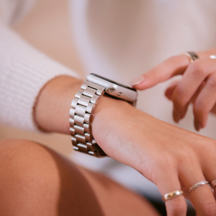 A close-up image of a person wearing Coconut Lane's Stainless Steel Apple Watch Strap - Silver, interacting with the device. The person is dressed in a light-colored sweater and adorned with several silver rings on their fingers. The blurred background accentuates the focus on the watch's elegant stainless steel band.