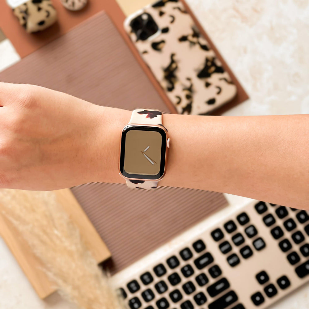 A person's wrist showcasing a Coconut Lane Milky Ivory Tort Apple Watch Strap featuring a stylish beige and black pattern. The background includes a keyboard and other accessories like a phone case and earbuds, all sharing the same chic beige and black design.