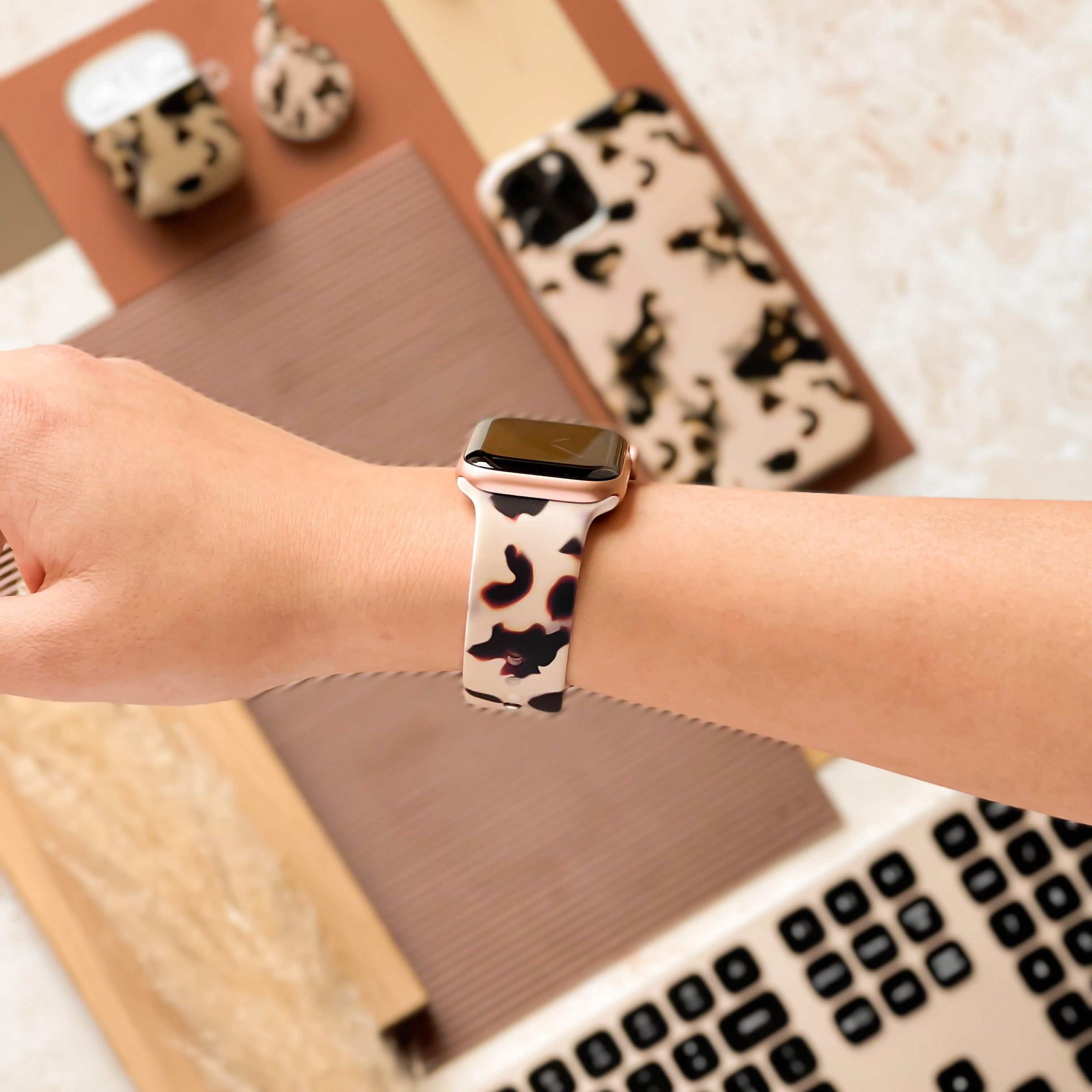 A person's wrist is shown wearing a Coconut Lane Milky Ivory Tort Apple Watch Strap. The background displays matching accessories, including a phone case, tablet case, keyboard, and other accessories, all featuring a similar animal print pattern.