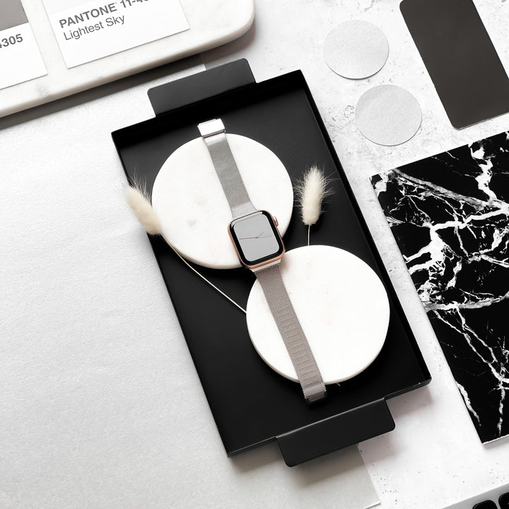 The Milanese Apple Watch Strap - Silver by Coconut Lane is showcased on a white circular stand set within a black tray. The tray is adorned with two white feathers. Nearby, Pantone color samples and marble-patterned papers highlight a minimalist design aesthetic.