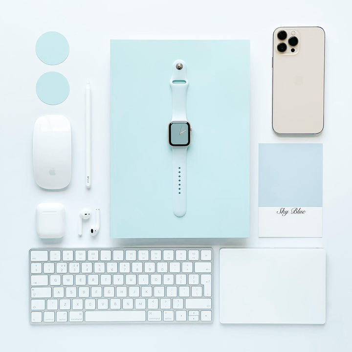 A neatly arranged flat lay featuring tech accessories in a minimalistic design. Includes an Apple Watch with a Coconut Lane NAKD Apple Watch Strap - Sky Blue, iPhone, keyboard, mouse, stylus, AirPods, two circular discs, and a card reading "Sky Blue," all set on a light blue rectangular base reflecting spring-inspired pastels.
