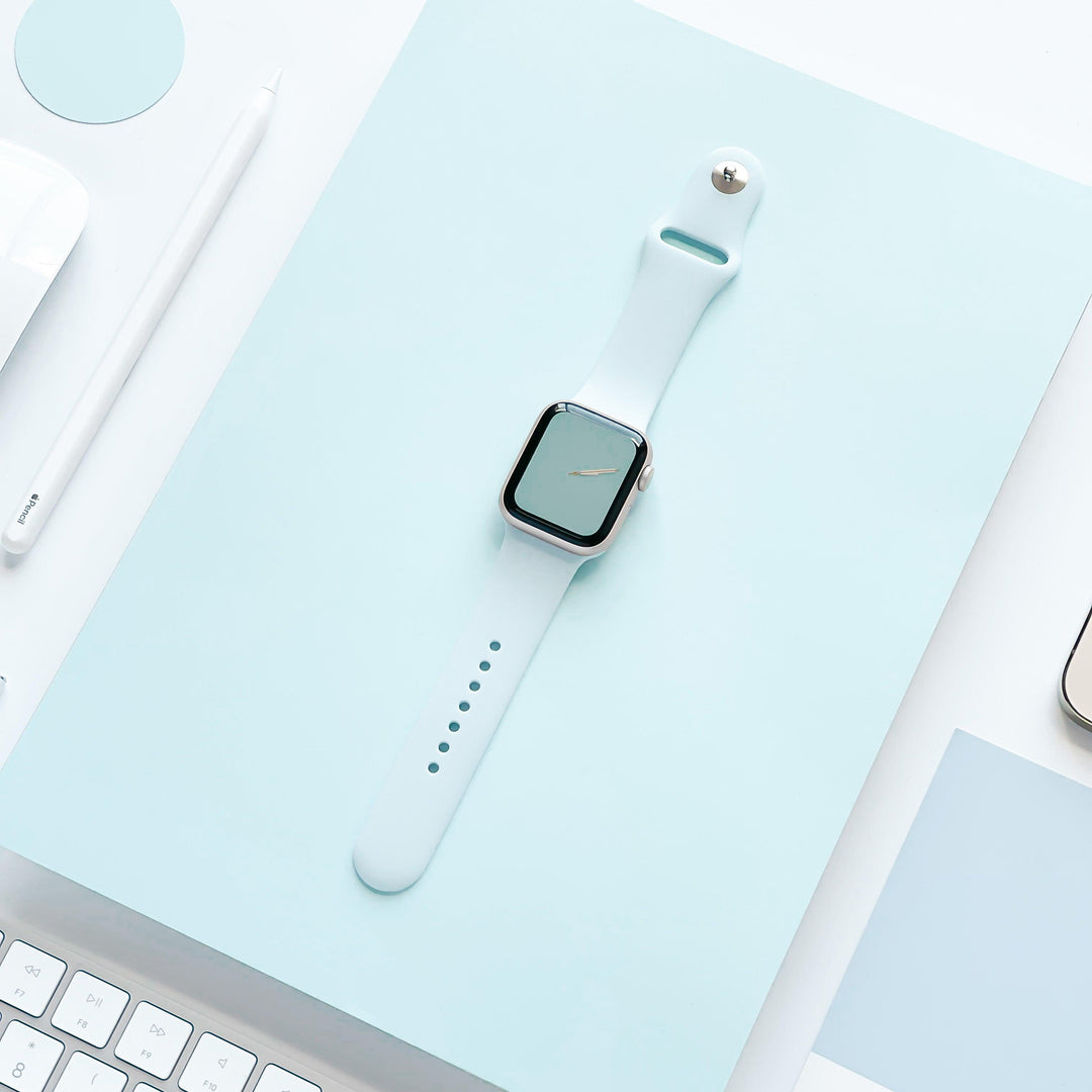 A light blue smartwatch with the NAKD Apple Watch Strap - Sky Blue by Coconut Lane is placed on a light blue surface. The smartwatch has a blank screen. Surrounding objects include a white keyboard, a white pen, and white paper, creating a minimalist, modern aesthetic with spring-inspired pastels.
