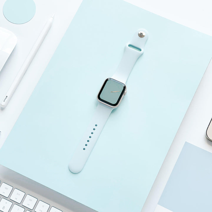 A light blue smartwatch with the NAKD Apple Watch Strap - Sky Blue by Coconut Lane is placed on a light blue surface. The smartwatch has a blank screen. Surrounding objects include a white keyboard, a white pen, and white paper, creating a minimalist, modern aesthetic with spring-inspired pastels.