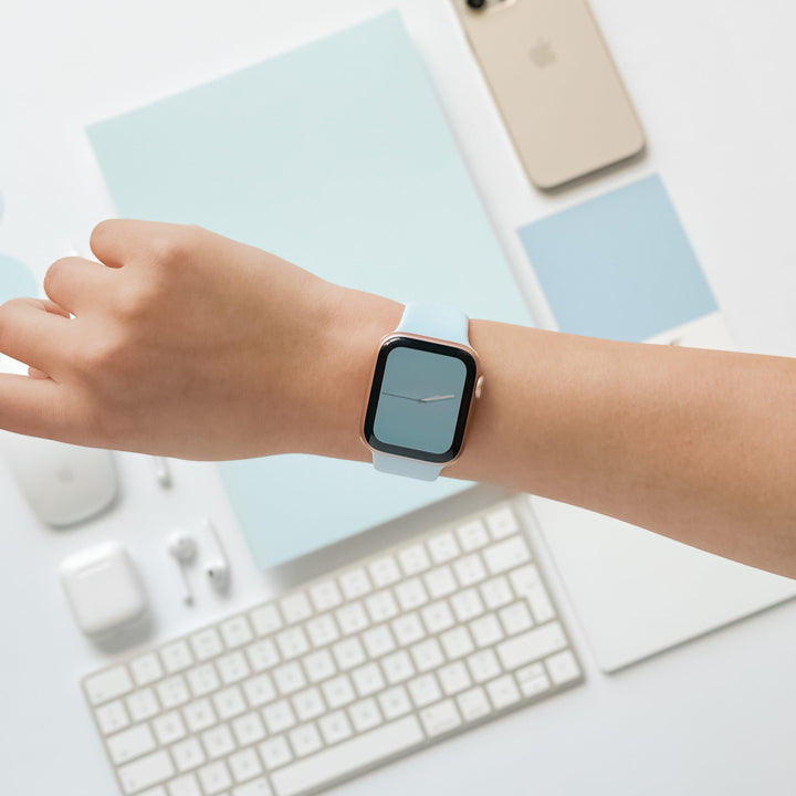 A hand wearing a spring-inspired pastel Apple Watch with the NAKD Apple Watch Strap - Sky Blue by Coconut Lane and a minimalist clock face, positioned above a desk with an iPhone, keyboard, AirPods, mouse, and small notebook, all color-coordinated in shades of blue and white.
