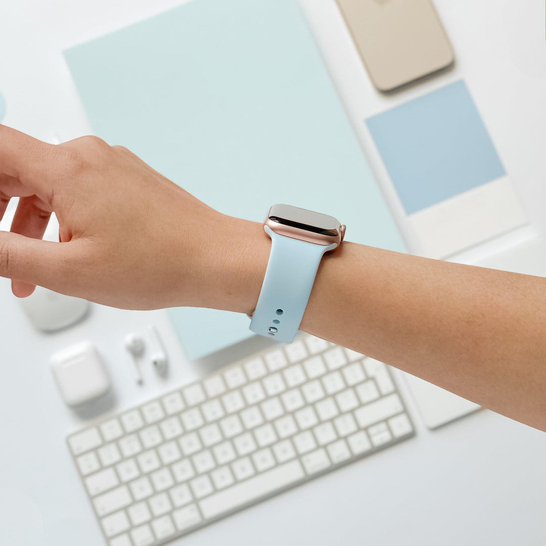 A person's arm is shown wearing the Coconut Lane NAKD Apple Watch Strap in Sky Blue. The background includes a white keyboard, wireless earbuds, and various sleek tech accessories in shades of white and spring-inspired pastels. The setting appears to be a clean and modern workspace.