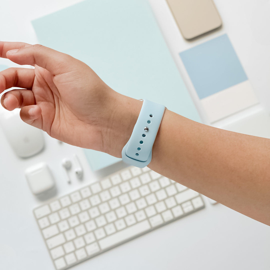Close-up of a person’s left wrist wearing a Coconut Lane NAKD Apple Watch Strap in Sky Blue. In the background, there’s a wireless mouse, keyboard, earbuds, phone, and other spring-inspired pastel tech accessories arranged neatly on a white surface.