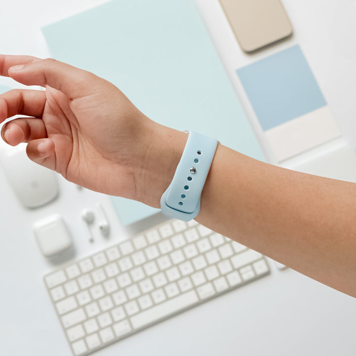 Close-up of a person’s left wrist wearing a Coconut Lane NAKD Apple Watch Strap in Sky Blue. In the background, there’s a wireless mouse, keyboard, earbuds, phone, and other spring-inspired pastel tech accessories arranged neatly on a white surface.