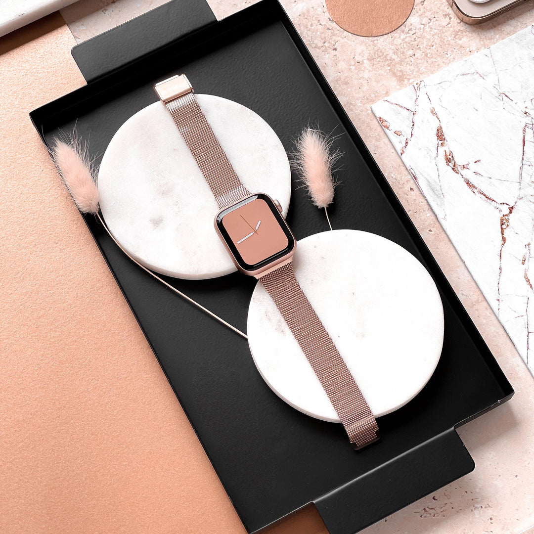 A rose gold smartwatch, featuring the Milanese Apple Watch Strap - Rose Gold by Coconut Lane, is neatly placed on two white circular bases within a black tray. Minimalist decorations, including beige feathers and marble-patterned items, surround the tray on a blush-toned surface.