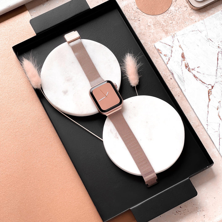 A rose gold smartwatch, featuring the Milanese Apple Watch Strap - Rose Gold by Coconut Lane, is neatly placed on two white circular bases within a black tray. Minimalist decorations, including beige feathers and marble-patterned items, surround the tray on a blush-toned surface.