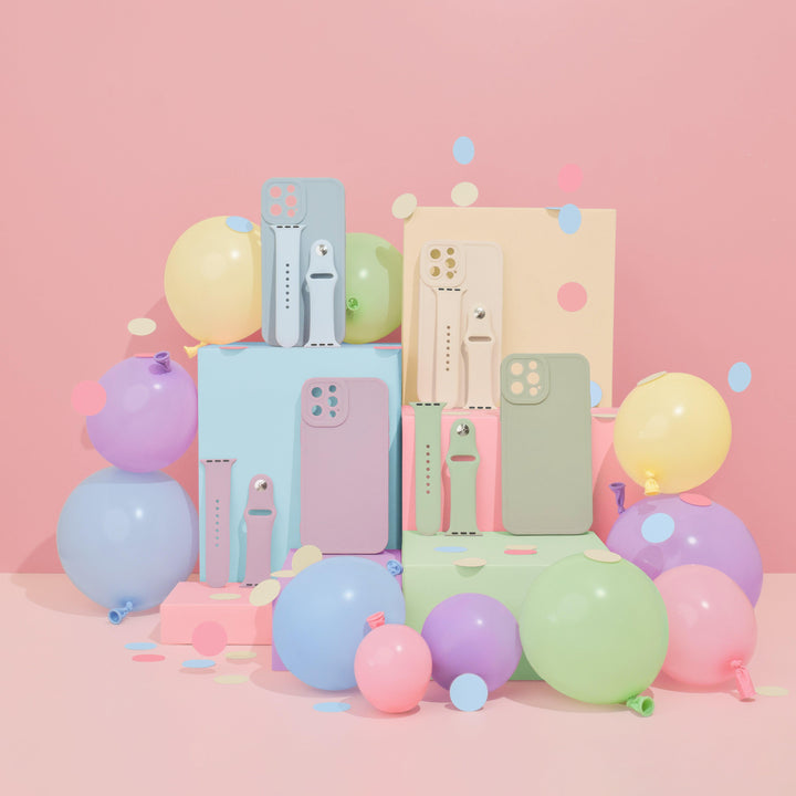 A pastel-themed arrangement features colorful smartphone cases and Coconut Lane's NAKD Apple Watch Straps in Sky Blue displayed on pastel blocks. The display is surrounded by balloons in blue, purple, yellow, green, and pink. Paper confetti is scattered across the scene, creating a festive, playful atmosphere.