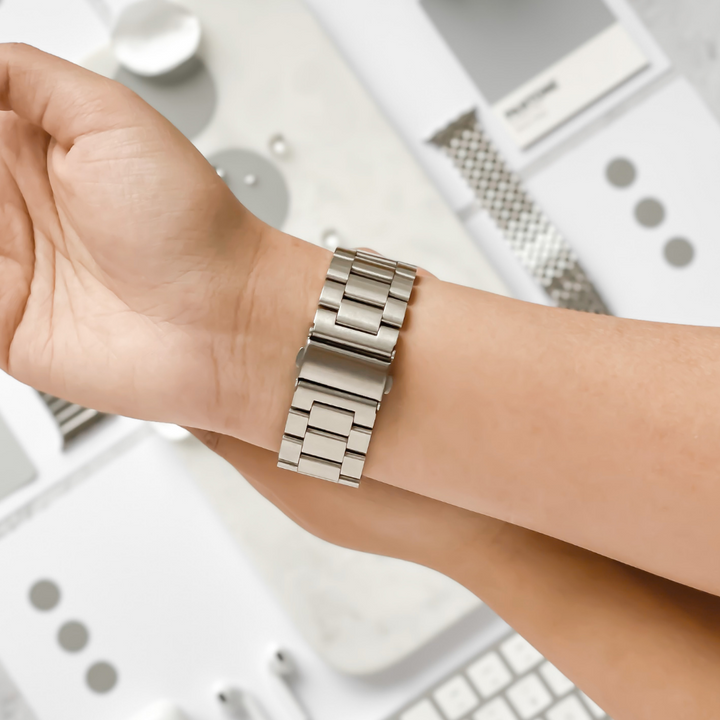 A hand wearing a sleek, Silver Stainless Steel Apple Watch Strap by Coconut Lane, with a blurred background featuring various watch components, tools, and materials in shades of gray and white.