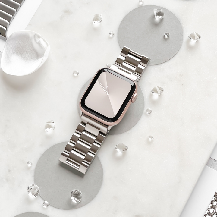 A sleek, silver smart watch with a Coconut Lane Stainless Steel Apple Watch Strap in Silver is elegantly displayed on a white marble surface. The watch is flanked by silver circles and scattered decorative crystal gems, creating a luxurious and modern aesthetic.