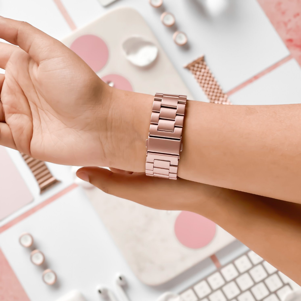 A wrist adorned with a blush-colored stainless steel Apple Watch strap by Coconut Lane is shown in close-up against a backdrop of light pink and white office supplies, including a keyboard, mouse, and decorative items. The adjustable strap features a sleek, modern design with interlocking links.