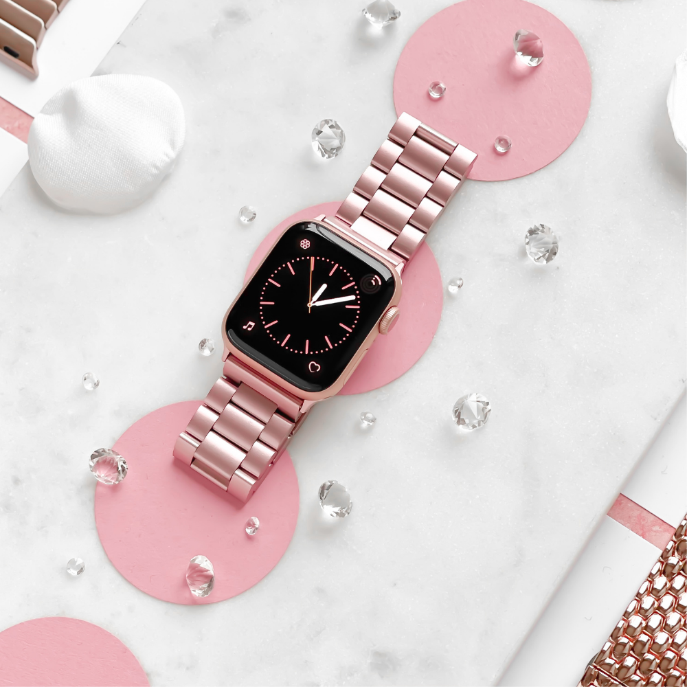 The Stainless Steel Apple Watch Strap - Blush by Coconut Lane is elegantly showcased on a marble surface adorned with pink polka dots. Encircling the watch are small clear crystals and white decorative elements, with the watch face indicating the time as approximately 10:09.